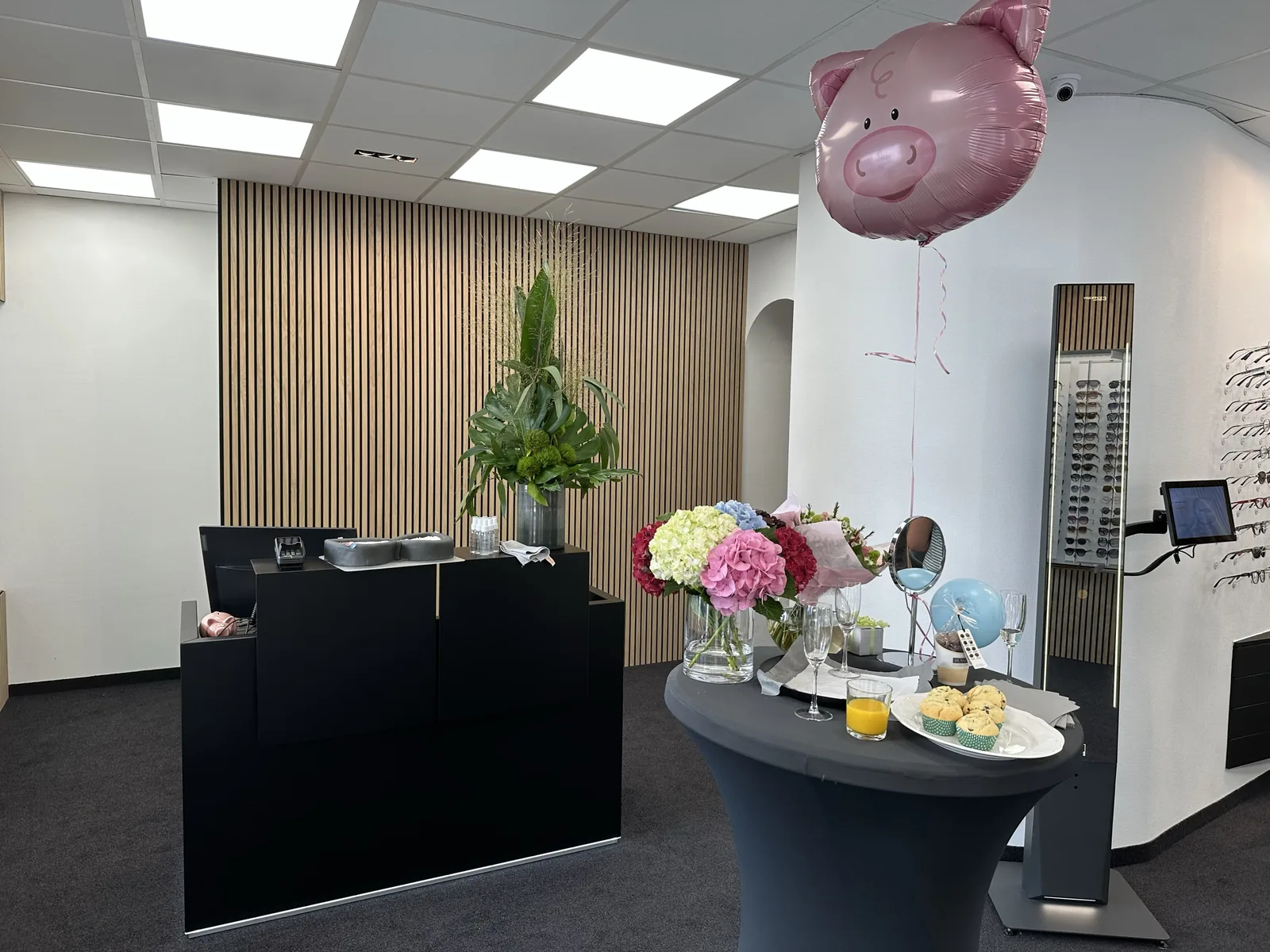 Fertiger Optiker-Laden am Eröffnungstag: Empfangstresen vor Holzlamellenwand, Blumen und Stehtisch