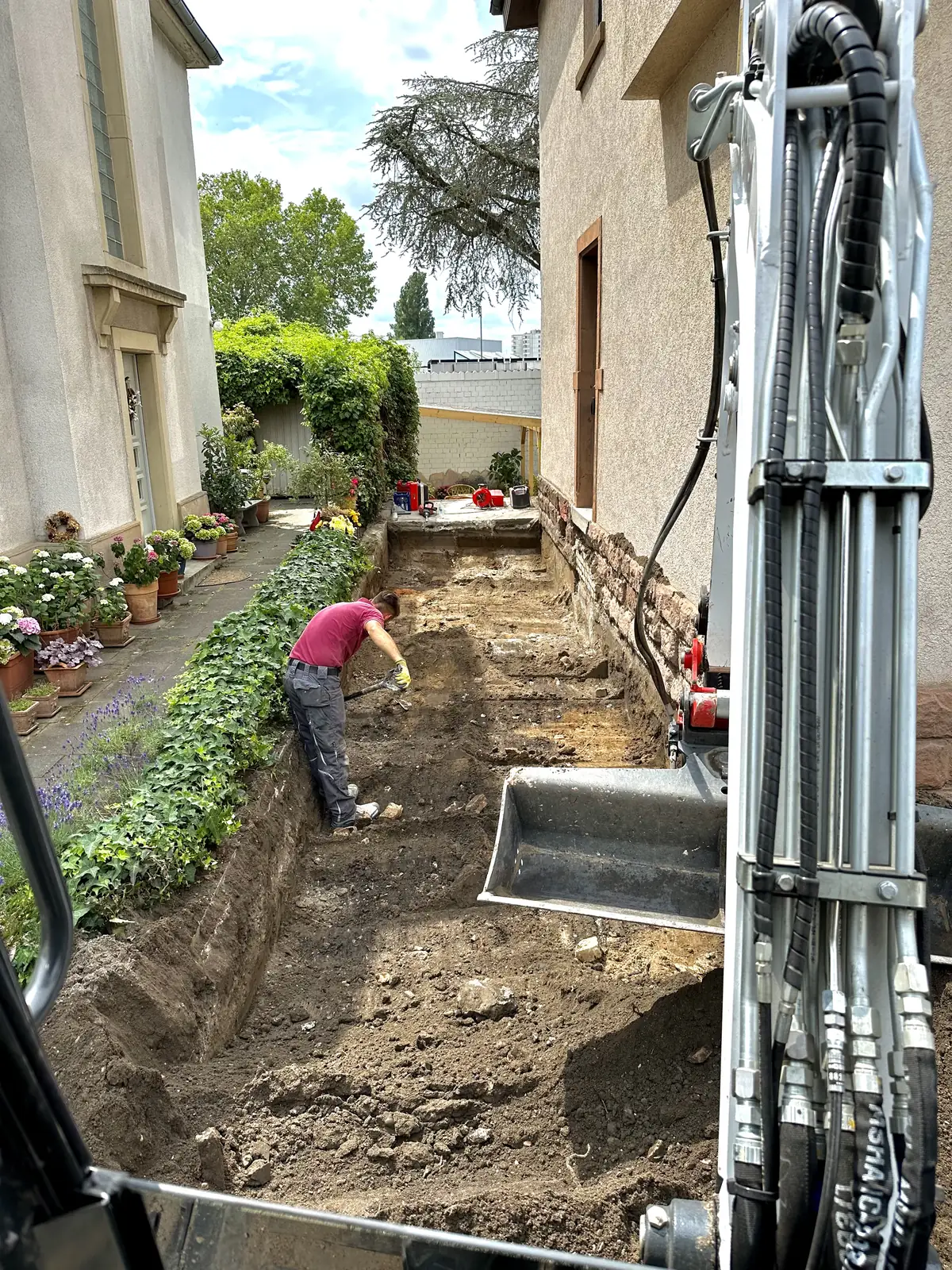 Baggerarm über dem ausgehobenen Graben neben dem Haus, Mitarbeiter richtet die Kante